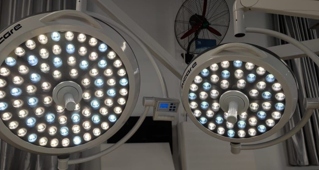 Ceiling-mounted dual LED surgical lights in an operating room, labeled “Aicare,” with multiple bright LED bulbs and control panels, used to provide high-intensity lighting during surgeries.